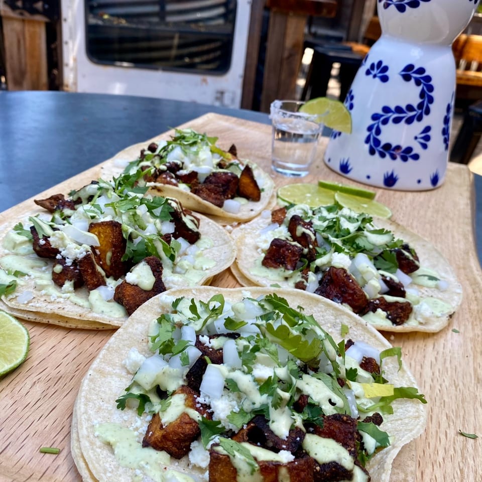 Four tacos with fried plantains, onions, cilantro, and green sauce, with a tequila shot.