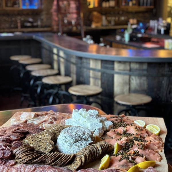 A charcuterie board with meats, cheeses, crackers, and lemons, in front of a dimly lit bar.