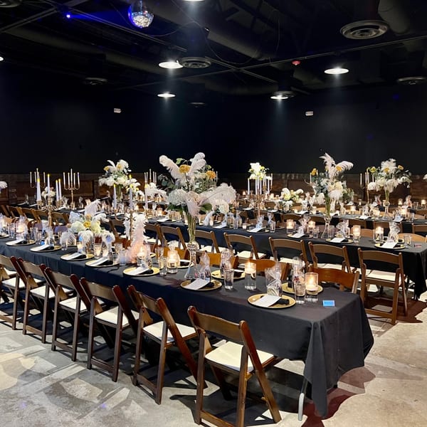 Elegant dining tables with black tablecloths, candles, and white floral centerpieces in a large event space.