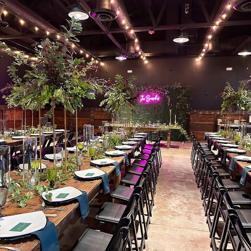 Long wooden tables set with green and white place settings, lush centerpieces, and string lights overhead.