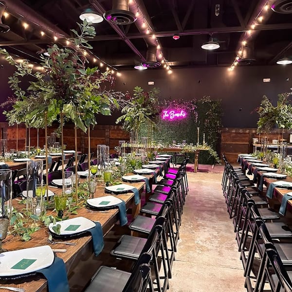 Long wooden tables set with green and white place settings, lush greenery, and string lights overhead.