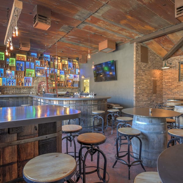 A rustic bar with a corrugated metal ceiling, brick walls, and a colorful backlit liquor display.