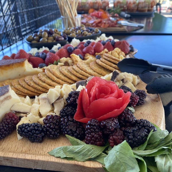 A wooden charcuterie board overflowing with cheeses, crackers, blackberries, and a tomato rose.