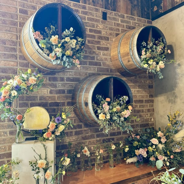 A brick wall decorated with three wine barrels overflowing with flowers, and a wooden counter with more floral arrangements.