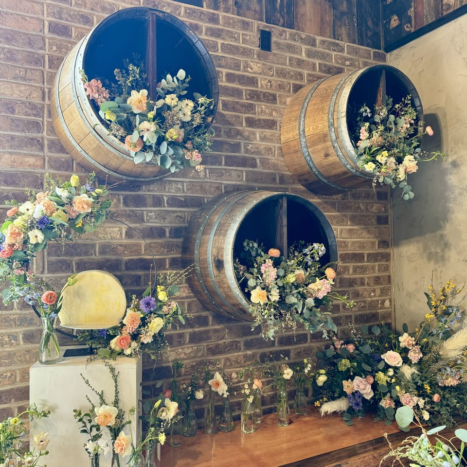 A brick wall decorated with three wine barrels overflowing with flowers, and a wooden counter with more floral arrangements.