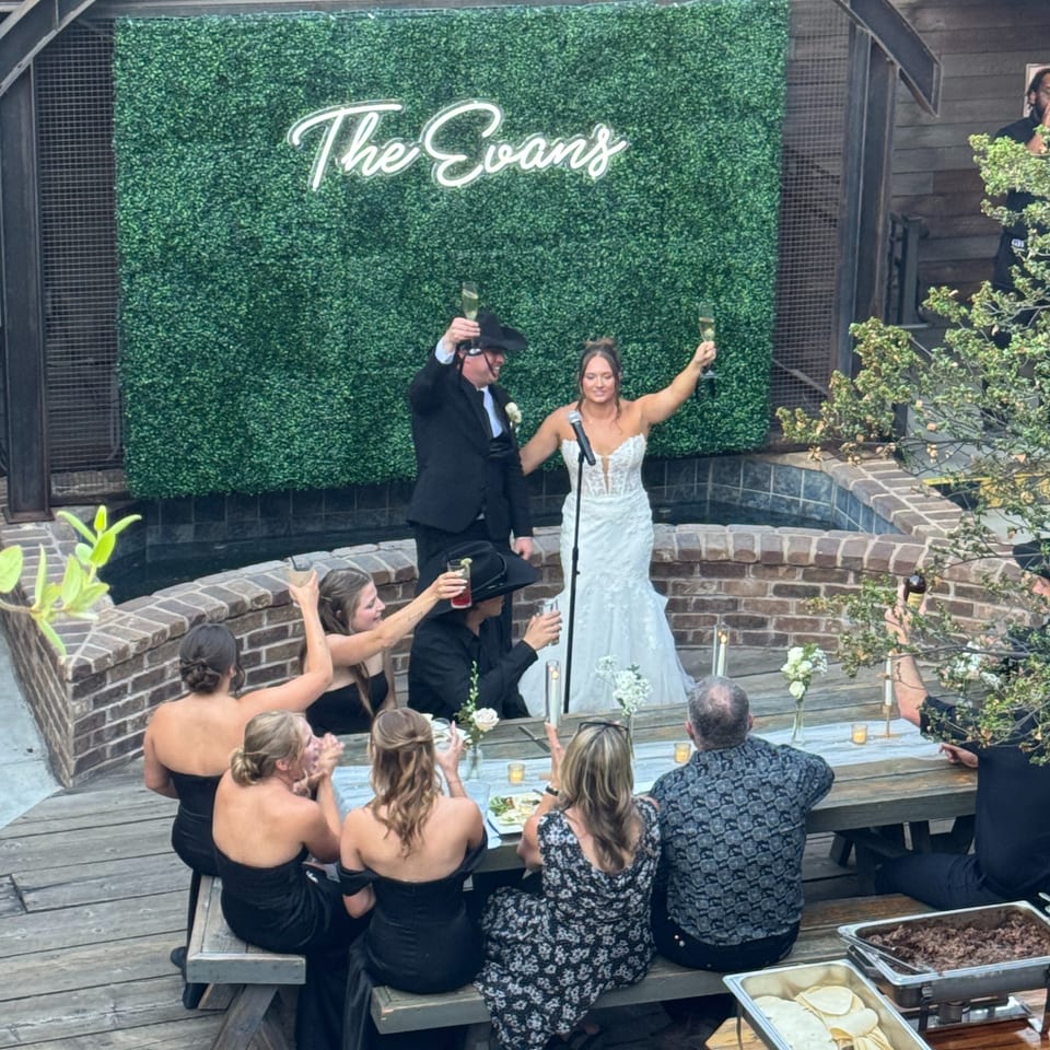 A wedding reception with a couple toasting in front of a green wall with "The Evans" in neon.