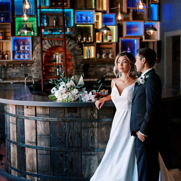 Bride and groom standing by a rustic wooden bar with colorful illuminated shelves in the background.