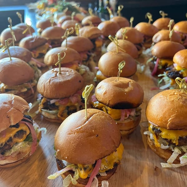 Numerous mini cheeseburger sliders with cheese, lettuce, and pickled onions on a wooden board.