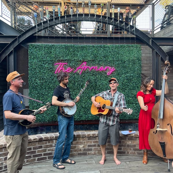 A band of four musicians playing string instruments on a patio with a neon "The Armory" sign.