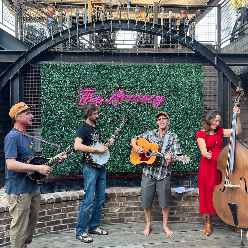 A band of four musicians playing string instruments on a patio with a neon "The Armory" sign.