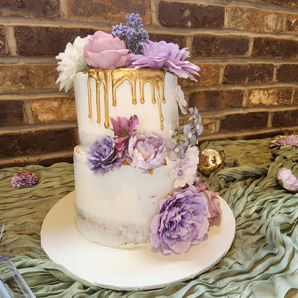 Two-tiered white cake with gold drip icing and purple, pink, and white floral decorations.
