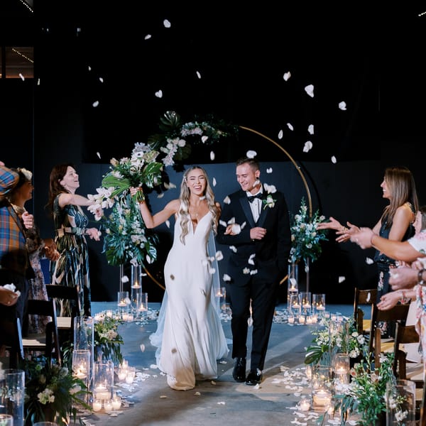 A happy bride and groom walk down an aisle showered with white petals and surrounded by candles.