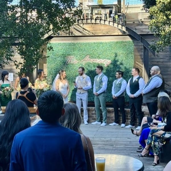 A wedding ceremony in an outdoor setting with a green wall backdrop and guests watching.