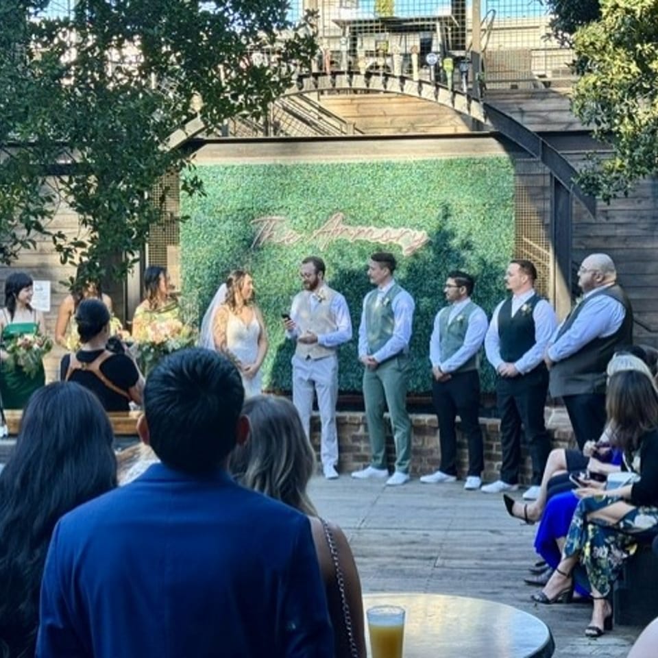 A wedding ceremony in an outdoor setting with a green wall backdrop and guests watching.