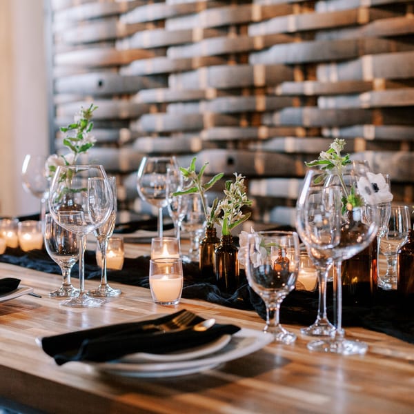 A long wooden table set with wine glasses, plates, black napkins, candles, and white flowers.