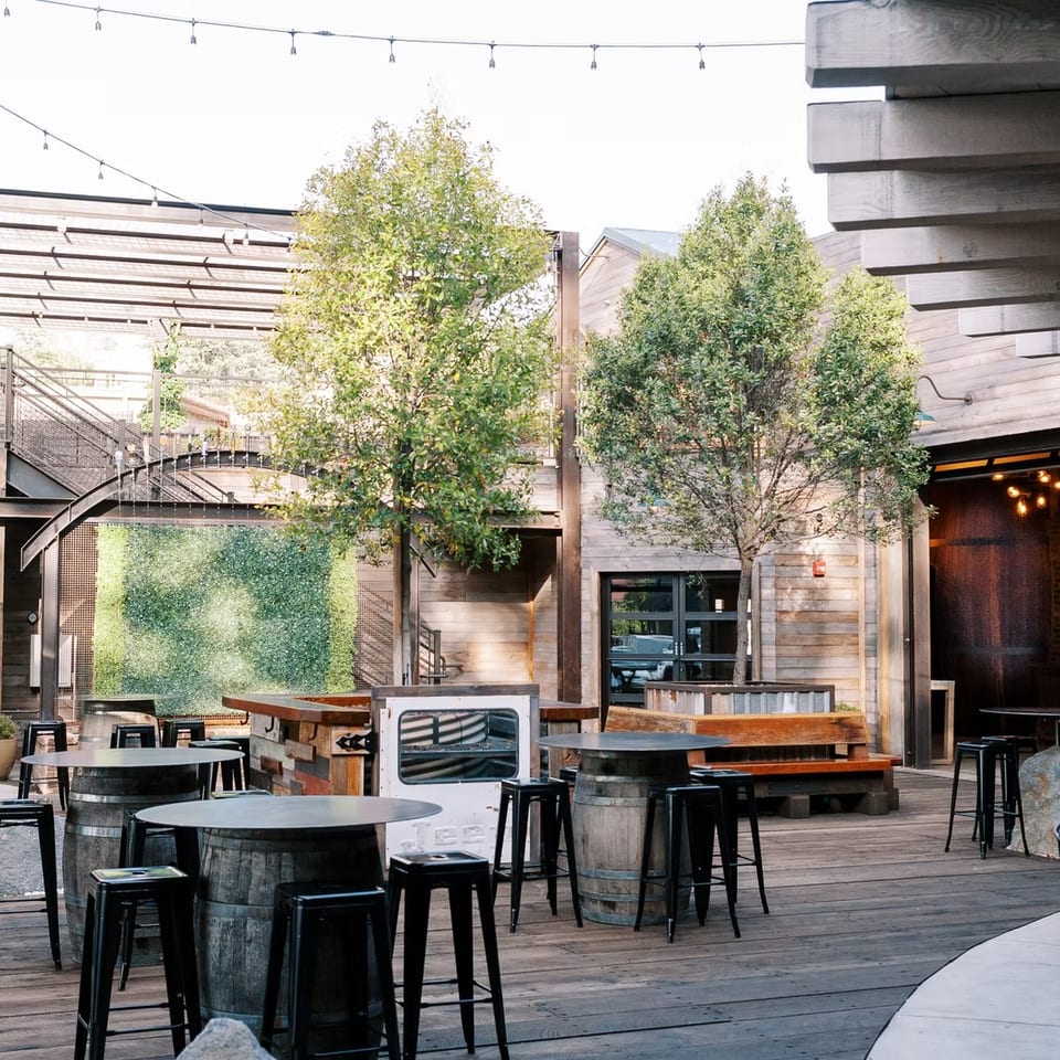 Outdoor patio with barrel tables, black stools, string lights, and wooden buildings with trees.