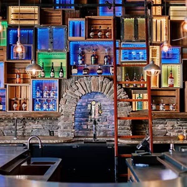 A rustic bar with a stone facade and wooden crates lit by colorful LED lights displaying bottles.