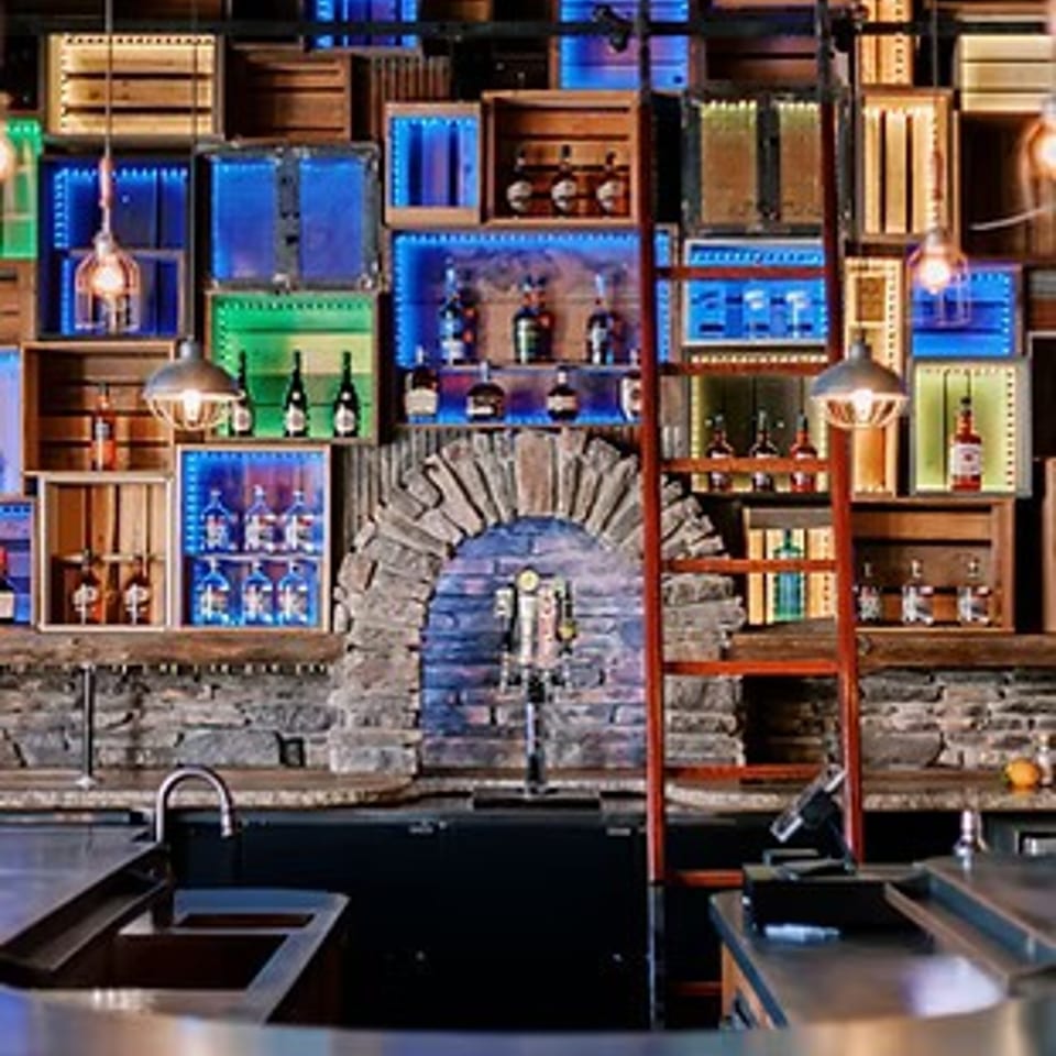 A rustic bar with a stone facade and wooden crates lit by colorful LED lights displaying bottles.