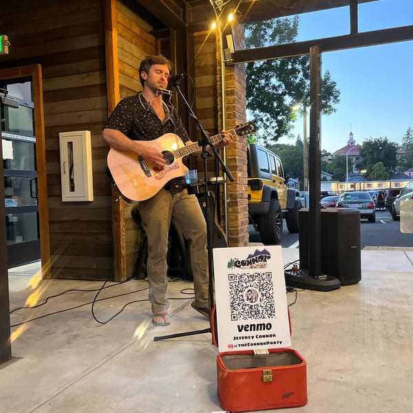 A man plays an acoustic guitar and harmonica outdoors, with a Venmo sign for tips nearby.