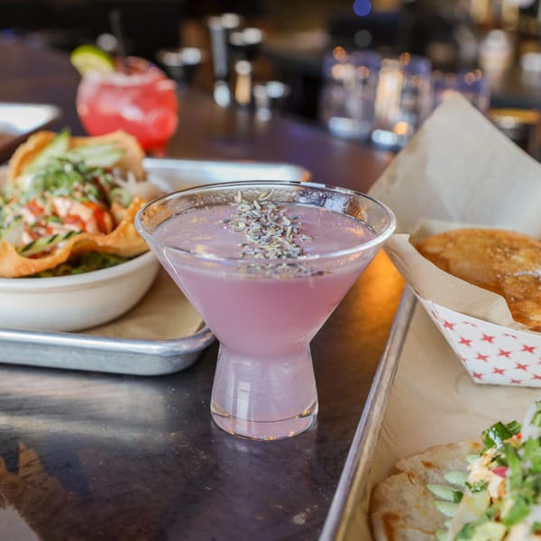 A lavender martini with food and drinks on a metal tray at a restaurant.