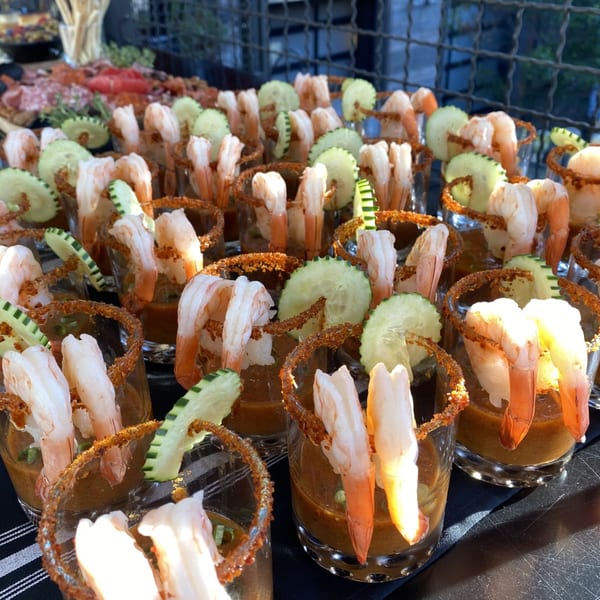 Numerous shrimp cocktail shooters with cucumber slices and chili-lime rims on a black tablecloth.
