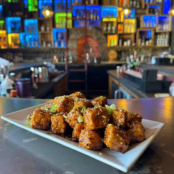 Crispy glazed tofu bites with sesame seeds and green onions on a white plate, against a colorful bar background.