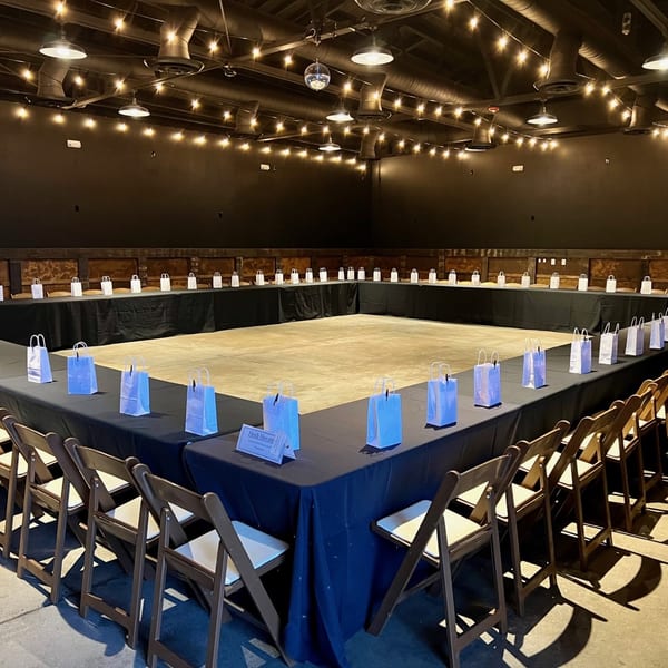 U-shaped conference table with black tablecloths, chairs, and white gift bags under string lights.