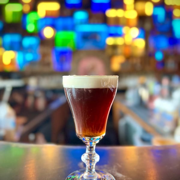 A glass of Irish coffee with a creamy white top sits on a dark bar with colorful lights in the background.