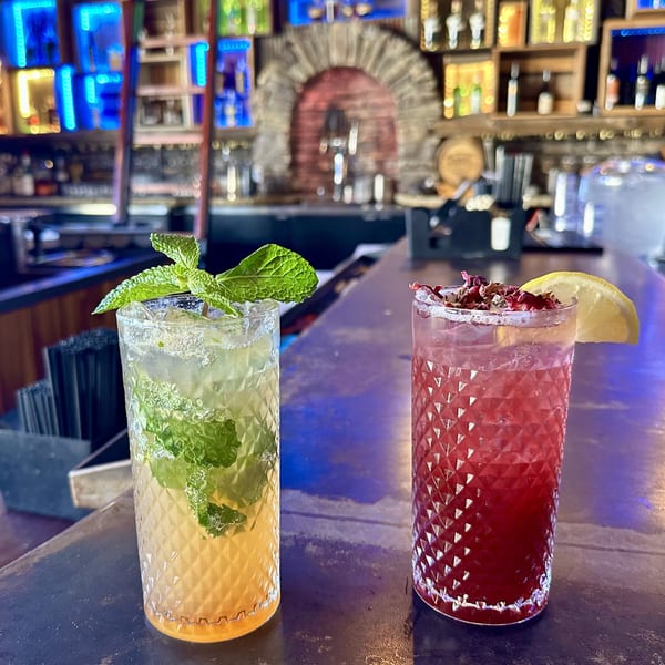 Two cocktails, one green with mint and one red with a lemon, on a bar with colorful lights.
