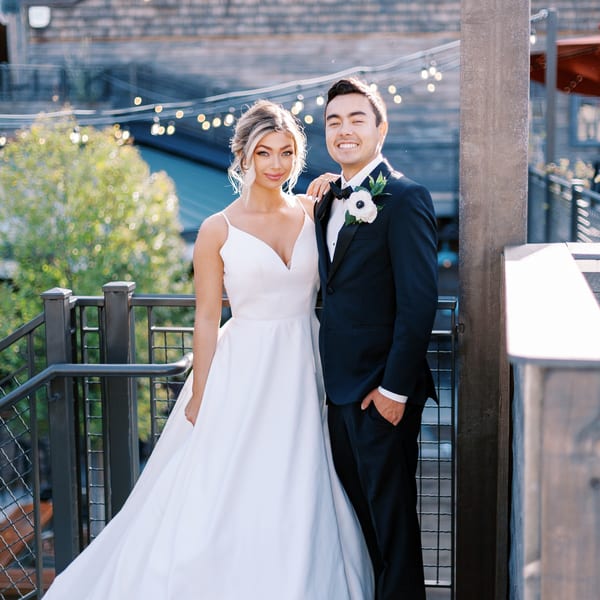 Smiling bride in white gown and groom in black tuxedo posing on a balcony with string lights.