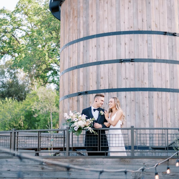 A smiling couple, the bride in white and groom in a tuxedo, stand on a balcony with a large wooden barrel structure behind them.