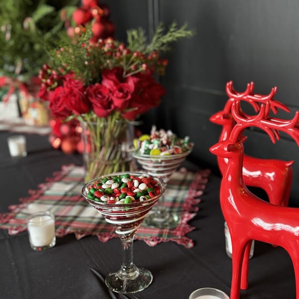 Christmas decorations with red roses, candy-filled glasses, and red reindeer figurines on a black tablecloth.