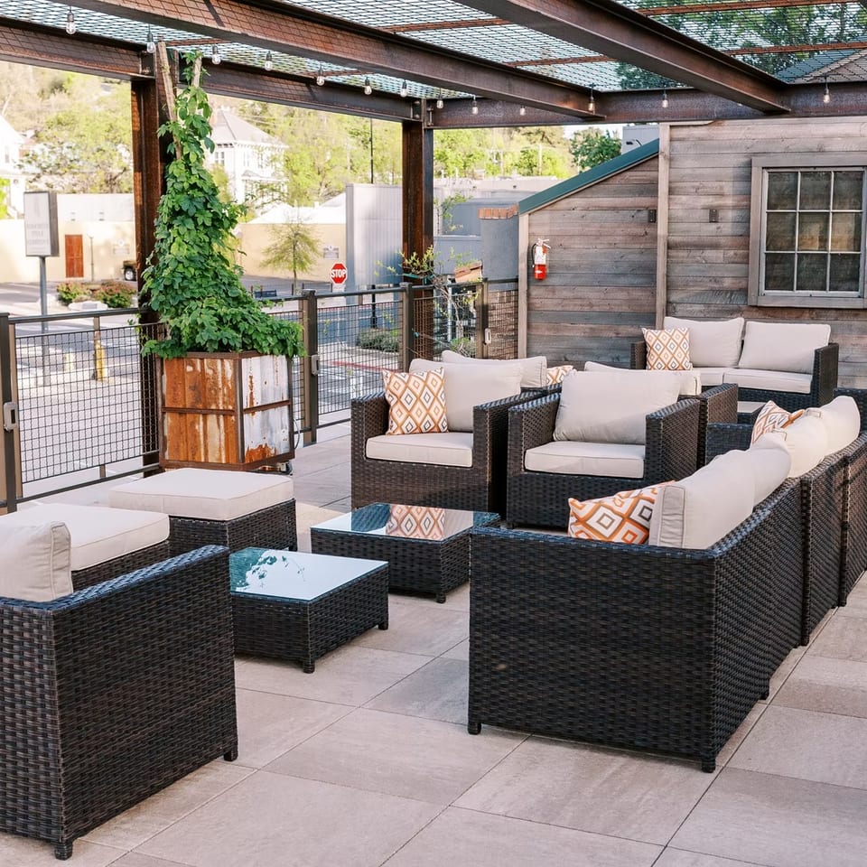 An outdoor patio with dark wicker seating, light cushions, and orange patterned pillows under a pergola.