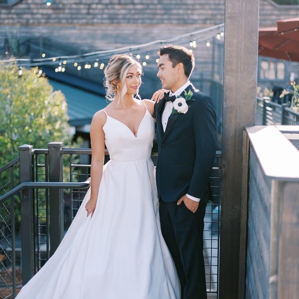 A newlywed couple poses on a balcony with string lights and a building in the background.