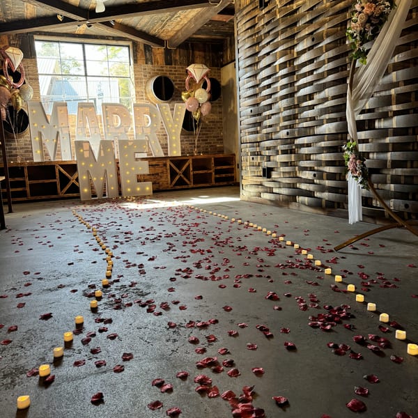 A room decorated for a proposal with "MARRY ME" lights, rose petals, and candles on the floor.