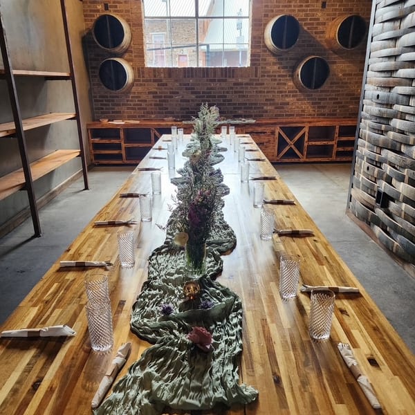 Long wooden dining table set with glasses and floral centerpieces in a rustic room with brick walls.