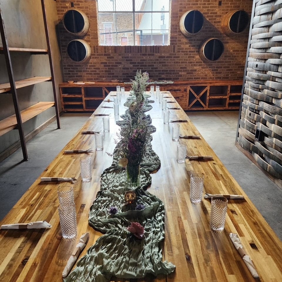 Long wooden dining table set with glasses and floral centerpieces in a rustic room with brick walls.