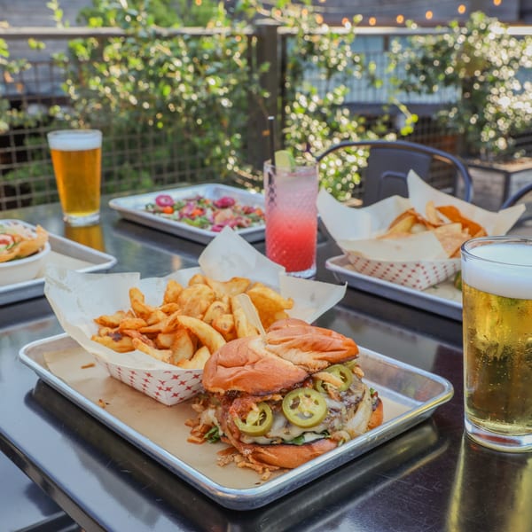 An outdoor table filled with burgers, fries, salads, and colorful drinks on a sunny patio.