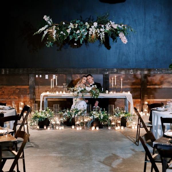 A couple sits at a candlelit head table adorned with white flowers and greenery at their wedding reception.