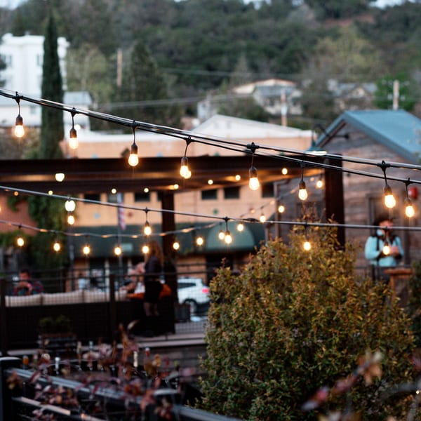 Outdoor patio with string lights, blurred people, and buildings in the background at dusk.
