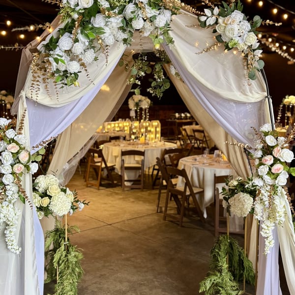 A wedding arch with white flowers and gold leaves leads to a reception area with round tables.