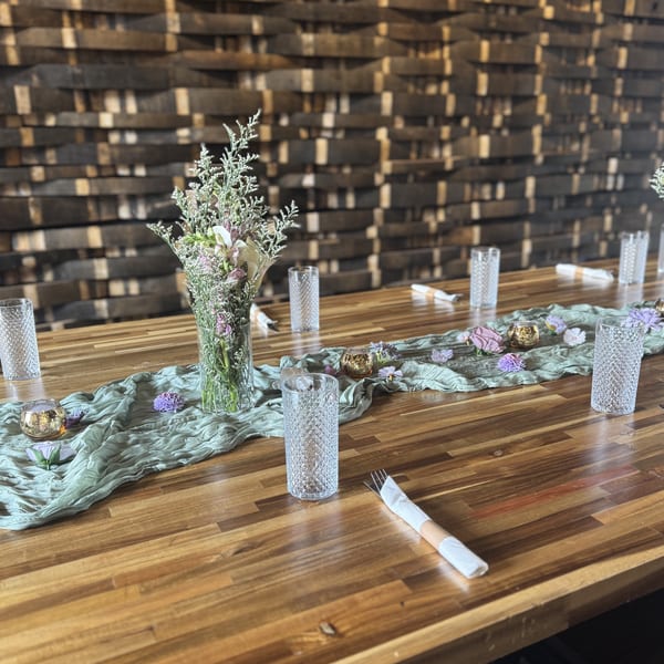 Long wooden table set with a green runner, floral centerpieces, glasses, and rolled silverware.