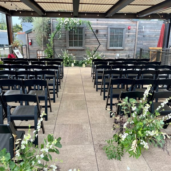 Outdoor wedding ceremony setup with black chairs, floral arch, and greenery on a patio.