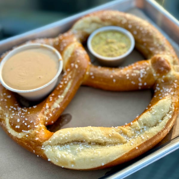 A large, salted pretzel on a tray with two dipping sauces: cheese and mustard.