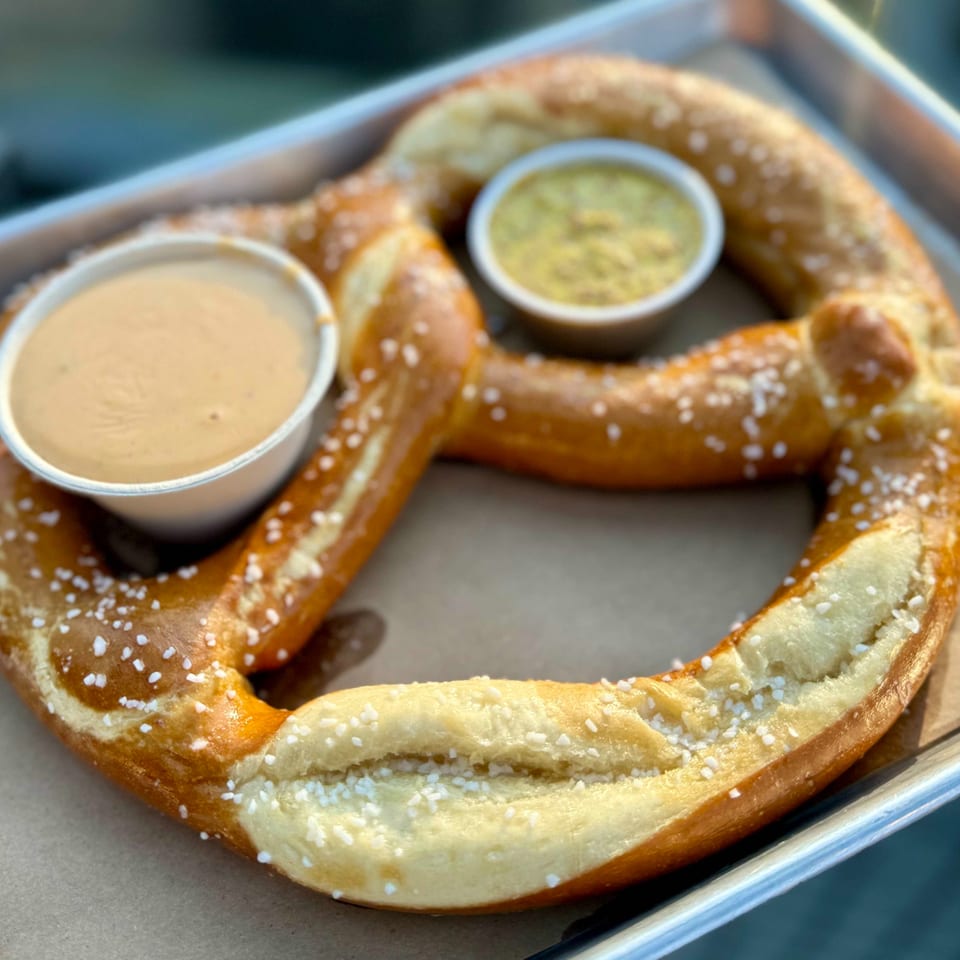 A large, salted pretzel on a tray with two dipping sauces: cheese and mustard.