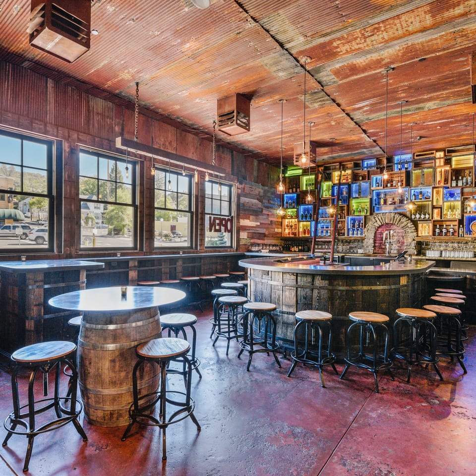 Rustic bar interior with a wooden barrel bar, colorful liquor display, and large windows.