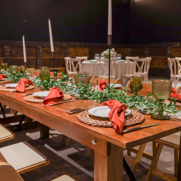 Long wooden dining table set with red napkins, green glasses, and candles, with round tables in the background.