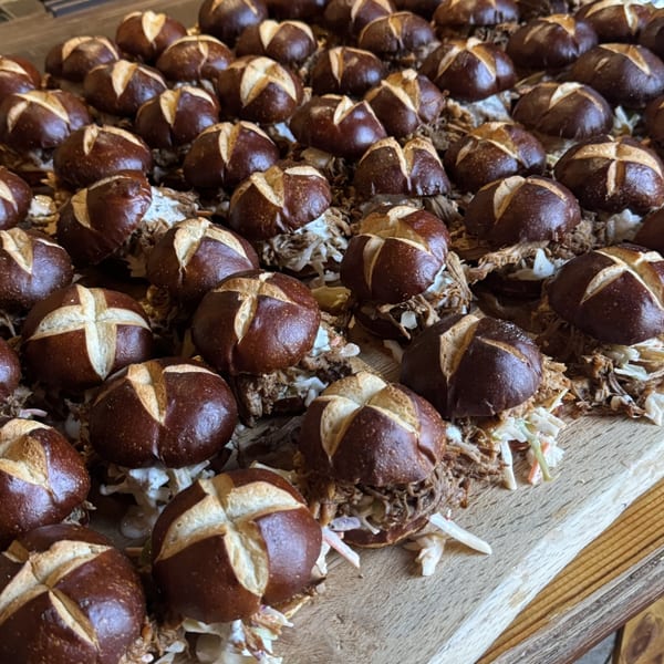 Numerous pulled pork sliders with coleslaw on pretzel buns arranged on a wooden board.